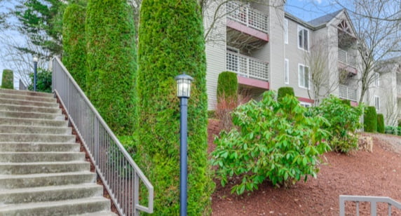 sidewalk view of residential building, stairs leading to building, surrounding landscaping, photo taken on a sunny day